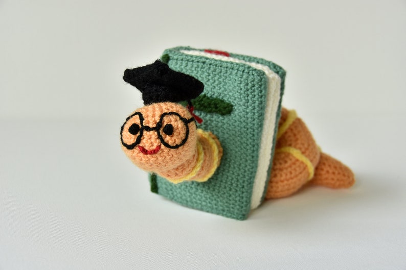 May include: A crocheted worm wearing glasses and a graduation cap, peeking out from behind a green book.