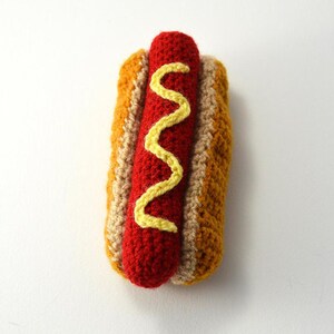 Op de afbeelding: Een gehaakte hotdog met mosterd, gemaakt om eruit te zien als een echte hotdog. De hotdog is rood, het broodje is bruin en de mosterd is geel.