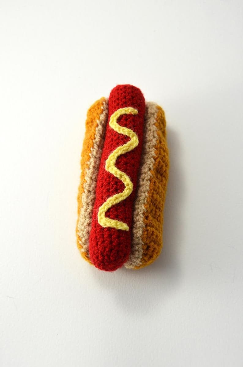 Op de afbeelding: Een gehaakte hotdog met mosterd, gemaakt om eruit te zien als een echte hotdog. De hotdog is rood, het broodje is bruin en de mosterd is geel.