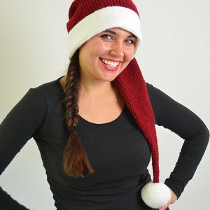 May include: A woman wearing a red and white crocheted Santa hat with a long white pom-pom. The hat has a long, red crocheted tail.