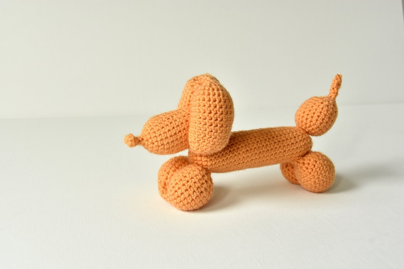 May include: A crocheted orange balloon dog toy. The dog is in a sitting position with its head turned to the side.