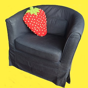 Op de afbeelding: Een rood en groen gehaakt aardbeienkussen op een zwarte fauteuil.