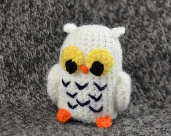 May include: A white crocheted owl with yellow and black eyes and orange feet. The owl is sitting on a gray fuzzy surface.