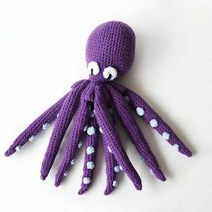 May include: A handmade purple crochet octopus with white and light blue accents. The octopus has eight long tentacles with light blue polka dots. The eyes are white with black pupils. The toy is made of soft yarn.