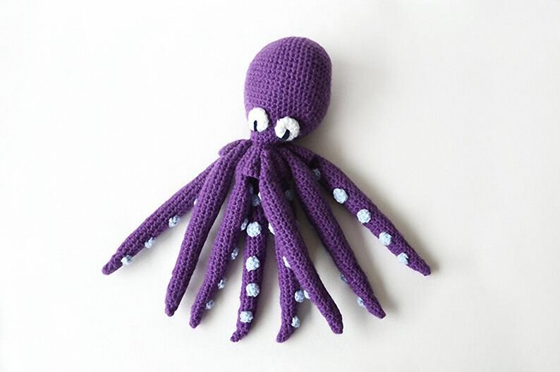 May include: A handmade purple crochet octopus with white and light blue accents. The octopus has eight long tentacles with light blue polka dots. The eyes are white with black pupils. The toy is made of soft yarn.