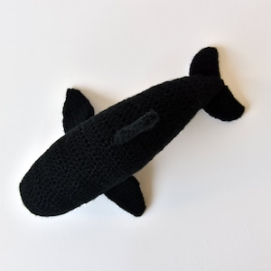 May include: Black crocheted stuffed whale toy. The whale is lying on its side with its tail in the air.