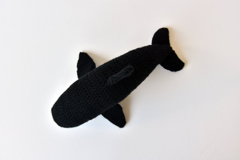 May include: Black crocheted stuffed whale toy. The whale is lying on its side with its tail in the air.