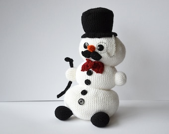 Può includere: Un pupazzo di neve bianco all'uncinetto con un cappello a cilindro nero, un papillon rosso, un baffo nero e occhi a bottone neri. Il pupazzo di neve tiene un bastone nero nella mano destra.