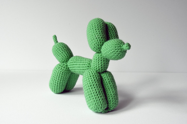 May include: A handmade, green crochet balloon dog sculpture. The dog is crafted with a textured stitch, featuring a rounded head, body, and legs, and a small tail. The sculpture is displayed against a plain white background.