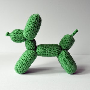 May include: A handmade green crochet balloon dog. The dog is crafted with a textured stitch, featuring a rounded body, legs, and head, with elongated balloon-like shapes. The dog is a vibrant green color and is set against a plain white background.