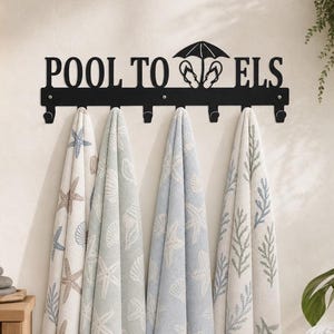 May include: A black metal wall-mounted towel rack featuring the text POOL TOWELS with a graphic of an umbrella and flip-flops. The rack has six hooks, four of which hold hanging towels decorated with blue and beige starfish and seashell patterns.