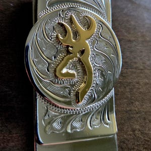 May include: Silver-toned metal money clip with a gold-colored deer emblem. The clip features an engraved floral design around the emblem and along the edges, creating a detailed and decorative accessory.