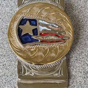 Puede incluir: Un clip de dinero plateado con un diseño grabado y ornamentado. El clip presenta un emblema circular dorado con una estrella azul y rayas rojas y blancas, que se asemeja a una bandera. El emblema está rodeado de intrincados adornos.