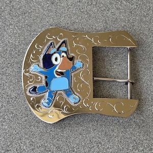 May include: A gold-coloured metal belt buckle featuring a cartoon dog character in shades of blue, black, and brown. The buckle has an ornate, swirling design around the character.