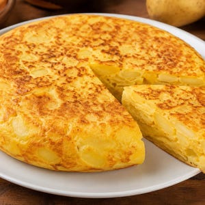 May include: A golden-brown Spanish tortilla, a thick omelet with potatoes and onions, sits on a white plate. A slice is cut and removed, revealing the potato filling. The dish is a traditional Spanish cuisine.