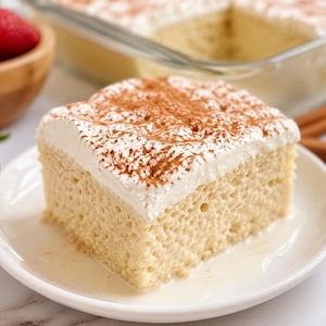Peut inclure: Une part de gâteau Tres Leches sur une assiette blanche, garnie de crème fouettée et saupoudrée de cannelle. Le gâteau est dans un bain de lait, avec un plat en verre et des fraises en arrière-plan. Un dessert savoureux.