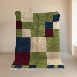 Könnte beinhalten: Ein rechteckiger Teppich mit geometrischem Muster. Der Teppich hat Quadrate in Grün, Beige, Marineblau und Burgunderrot. Der Teppich hat einen Fransenbesatz am unteren Rand. Der Teppich wird vor einem neutralen Hintergrund präsentiert.