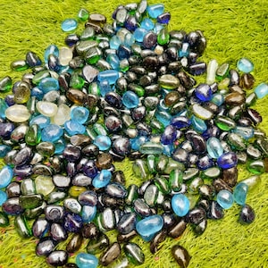 Puede incluir: Un conjunto de guijarros de vidrio pulido en varios colores, incluyendo azul, verde y negro, sobre una superficie verde. Los guijarros tienen una forma suave y redondeada y reflejan la luz. Ideal para fines decorativos.