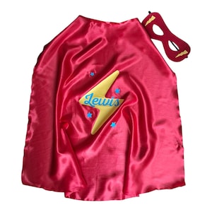 May include: A shiny red satin superhero cape featuring a large yellow lightning bolt emblem with the name Lewis written in blue script across the centre, accented by small blue stars, accompanied by a matching red felt eye mask with gold lightning bolts.