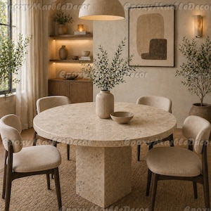 Puede incluir: Una mesa de comedor redonda con una superficie moteada de color claro, sostenida por una base cuadrada. Cuatro sillas beige tapizadas con patas de madera oscura rodean la mesa. Una lámpara colgante beige cuelga encima, en una habitación con tonos neutros.