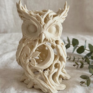 Könnte beinhalten: Eine cremeweiße Eulenfigur mit einem komplizierten, durchbrochenen Design aus ineinander verschlungenen Zweigen, Blättern und einem Halbmond in der Mitte der Brust. Die Skulptur hat eine matte Oberfläche und detaillierte Federstrukturen.