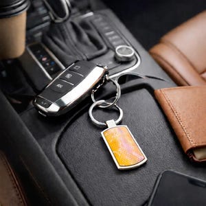 Peut inclure: Un porte-clés en métal argenté avec une étiquette rectangulaire au motif texturé dégradé orange et rose rappelant la peau de serpent, attaché à une clé de voiture noire et argentée avec trois boutons et une bordure métallique.