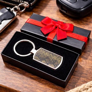 May include: A rectangular metal keyring featuring a dark brown and black snakeskin pattern inlay, attached to a circular silver ring. The keyring is presented inside a black gift box adorned with a vibrant red ribbon tied in a bow.