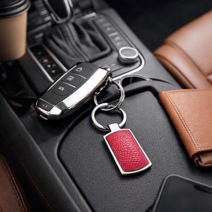 May include: A rectangular keychain featuring a textured, bright red leather-like inlay framed in polished silver-tone metal, attached to a silver metal split ring and a black car key fob with three buttons and a metallic silver trim.