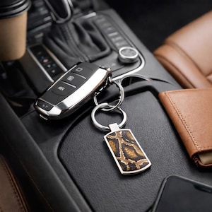 May include: A rectangular metal keyring featuring a brown and black snakeskin pattern inlay, attached to a silver-toned split ring. The keyring is positioned next to a black car key fob with silver accents on a dark textured console surface.