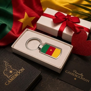 Könnte beinhalten: Ein Schlüsselanhänger mit der Flagge Kameruns, präsentiert in einer Geschenkbox mit roter Schleife. Der Schlüsselanhänger hat ein grünes, rotes und gelbes Design mit einem Stern. Eine Karte trägt die Aufschrift "Proudly Cameroonian".