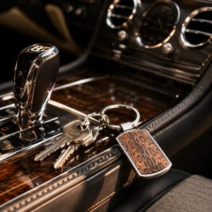 May include: A metal keyring featuring a rectangular tag with a brown and black snakeskin pattern, attached to a silver split ring holding two silver keys. The keyring rests on a dark wood and chrome centre console near a gear shift lever.