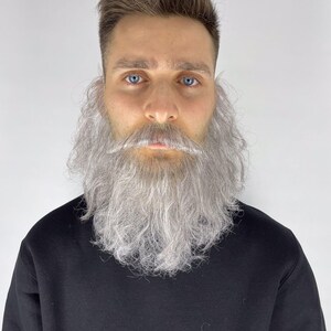 May include: A man with blue eyes and light brown hair is wearing a black sweatshirt. He has a long, gray beard and mustache. The beard is styled to frame the face.