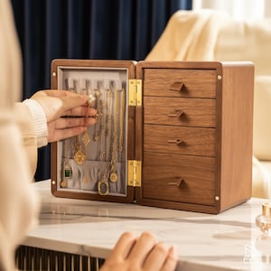 May include: A wooden jewelry box with a hinged door and four drawers. The open door reveals necklaces hanging on a gray fabric. The box is made of brown wood with brass-colored hardware. The jewelry includes gold necklaces with various pendants.