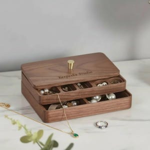 Puede incluir: Una caja de joyería de madera de dos niveles, llena de anillos, collares y pendientes. La caja es de madera oscura y tiene un pomo dorado en la tapa. El texto "Keepsake Studio" está grabado en la tapa.