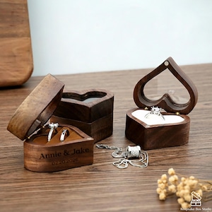 May include: Heart-shaped wooden ring boxes with clear lids and velvet interiors. One box is open, displaying two rings, and engraved with "Annie & Jake" and a date. Another box holds a single ring. A silver necklace lies nearby.
