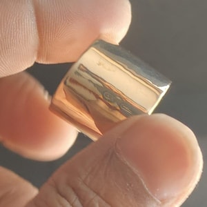 May include: A gold-colored ring with a rectangular design. The ring is held between fingers, showcasing its polished surface and simple, elegant shape. The ring's smooth, reflective surface catches the light, highlighting its metallic sheen.