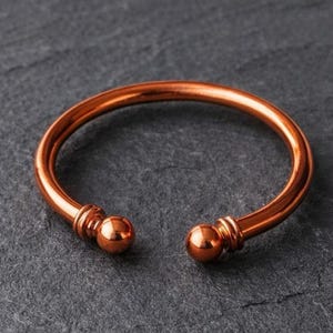 May include: A copper-colored open cuff bracelet with rounded terminals. The bracelet has a smooth, polished finish and a simple, elegant design. The terminals are spherical and add a decorative element to the bracelet.