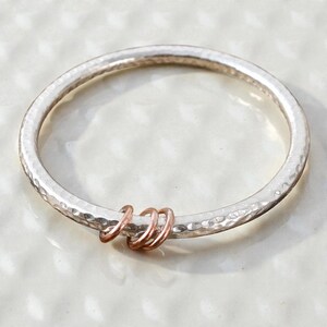 Könnte beinhalten: Ein silbernes Armreif-Armband mit gehämmertem Finish und drei roségoldenen Ringen. Das Armband hat eine strukturierte Oberfläche und ein kreisförmiges Design. Die roségoldenen Ringe sind klein und setzen einen farblichen Akzent.