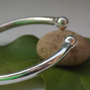 Könnte beinhalten: Ein offenes, silberfarbenes Armreif-Armband mit abgerundeten Enden. Das Armband liegt auf einem grünen Blatt und einem kleinen, runden, hellbraunen Stein. Die polierte Oberfläche reflektiert das Licht.