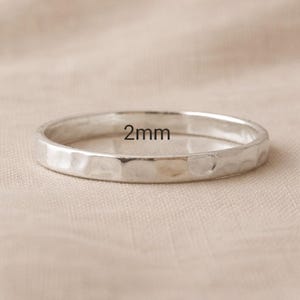 May include: A silver ring with a hammered texture. The ring is 2mm wide and sits on a neutral-colored surface. The ring is a simple band design.