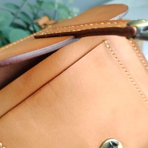 May include: Close-up of a tan leather wallet with a flap closure. The wallet features visible stitching along the edges and a brass-colored clasp. The leather has a smooth texture and a natural color.