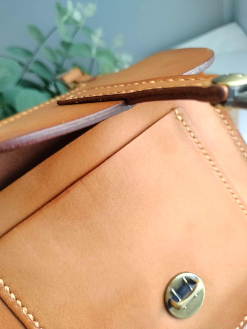 May include: Close-up of a tan leather wallet with a flap closure. The wallet features visible stitching along the edges and a brass-colored clasp. The leather has a smooth texture and a natural color.