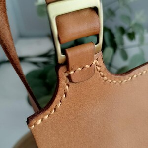 May include: Close-up of a tan leather bag strap with a gold-colored metal buckle. The leather is stitched with a contrasting light-colored thread. The bag strap is a light brown color.