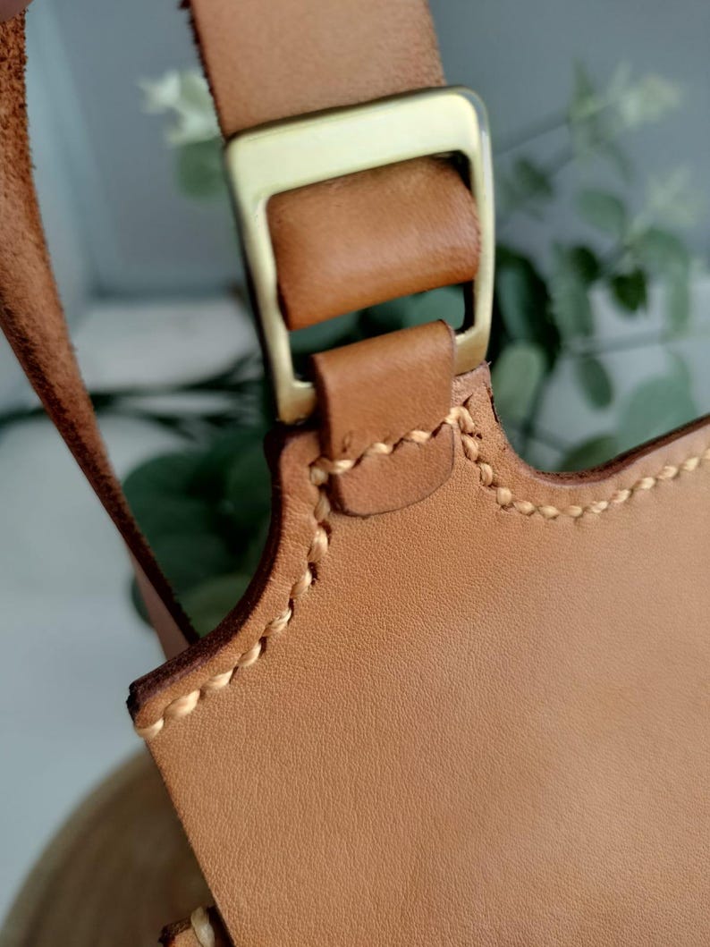 May include: Close-up of a tan leather bag strap with a gold-colored metal buckle. The leather is stitched with a contrasting light-colored thread. The bag strap is a light brown color.