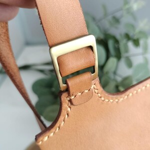 May include: Close-up of a tan leather bag strap with a gold-colored metal buckle. The bag's edge is visible, featuring detailed stitching. The leather has a natural, textured appearance.