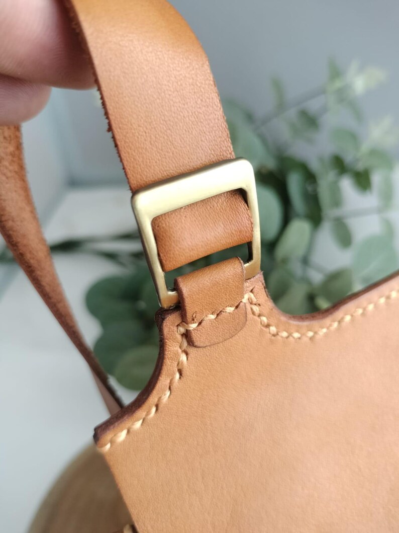 May include: Close-up of a tan leather bag strap with a gold-colored metal buckle. The bag's edge is visible, featuring detailed stitching. The leather has a natural, textured appearance.