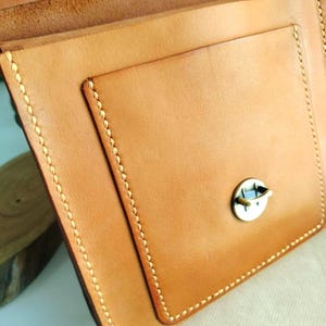 May include: Close-up of a handcrafted, light brown leather bag with a flap and a small pocket. The bag features visible stitching in a contrasting light color and a metal clasp. The leather has a natural, textured appearance.