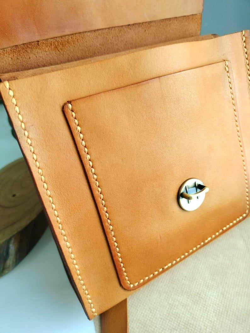 May include: Close-up of a handcrafted, light brown leather bag with a flap and a small pocket. The bag features visible stitching in a contrasting light color and a metal clasp. The leather has a natural, textured appearance.