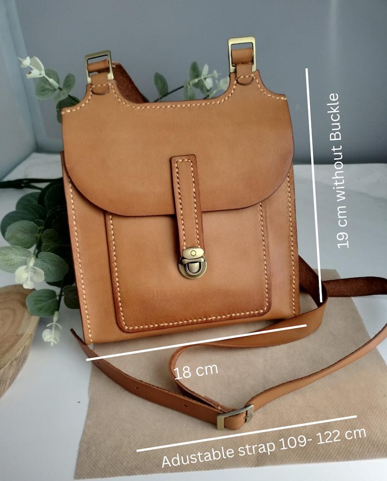 May include: A tan leather crossbody bag with an adjustable strap. The bag has a flap closure with a metal clasp and a small front pocket. The bag is 19 cm without the buckle and 18 cm wide. The adjustable strap is 42.9 to 48 inches.