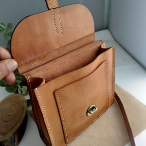 May include: A light brown leather crossbody bag with a flap closure and a front pocket. The bag has a long, adjustable strap and a metal clasp. The stitching is visible and the bag appears to be handmade.